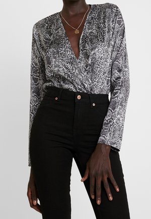 Persoon die zwarte hoge taille broek en een zwart-wit slangenprint overslag top met lange mouwen, V-hals en een ronde hangersketting draagt.