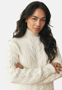 White cable-knit turtleneck sweater featuring ribbed accents, textured patterns, and long sleeves.