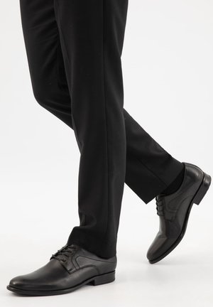 Persoon draagt zwarte nette schoenen en zwarte broek, met één voet lichtjes opgetild alsof die vooruitstapt.