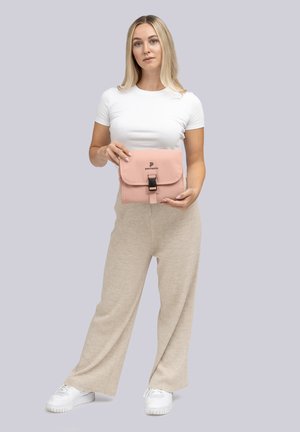 Femme aux cheveux blonds portant un haut blanc et un pantalon beige, tenant une pochette pliée rose avec un fermoir noir, étiquetée "pactastic."