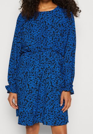Femme portant une robe longue à manches longues bleue et noire à motif floral, avec une taille cintrée et des poignets à volants, debout devant un fond uni.