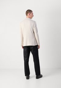 Blazer beige chiaro con tessuto testurizzato, con uno spacco posteriore singolo e due tasche laterali, abbinato a pantaloni neri e scarpe nere lisce.