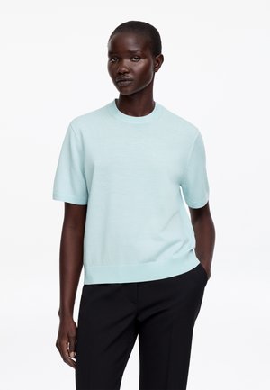 Kvinde med mørk hud og kort hår iført en lyseblå kortærmet striktop og sorte bukser, stående mod en hvid baggrund.