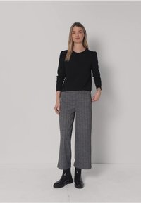 Schwarzes Langarmshirt mit rundem Ausschnitt, kombiniert mit grauen, weit geschnittenen Hosen im Hahnentrittmuster und schwarzen Stiefeletten.