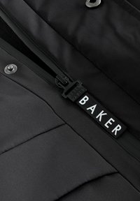 Melnā polsterētā jaka ar gludu tekstūru, kas aprīkota ar rāvējslēdzēju, spiedpogām un redzamu zīmola etiķeti ar vārdu "BAKER" baltā krāsā.