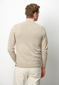 Suéter beige con cuello alto, mangas largas y puños acanalados. Textura suave y corte ajustado, combinado con pantalones de color claro.