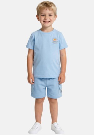 Jeune garçon souriant portant un t-shirt bleu clair et un short cargo avec des baskets blanches, debout devant un fond uni.