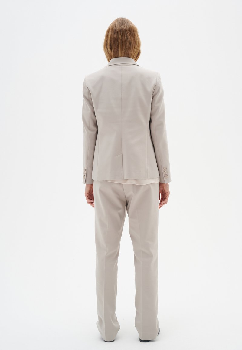 Hellgrauer Anzug, bestehend aus einem taillierten Blazer und gerade geschnittenen Hosen. Der Stoff wirkt glatt mit minimaler Detailierung und Knopfakzenten.
