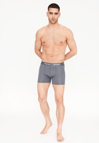 Boxers gris con una cinturilla negra que presenta texto blanco. Tejido suave con un diseño ajustado, costura delantera y longitud hasta medio muslo.