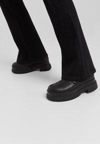 Mustat, tukevat platform-kengät, joissa on sileä nahkamainen pinta, pyöreä kärki ja teksturoitu pohja, yhdistettyinä mustiin leveälahkeisiin housuihin.