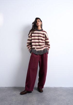 Kvinde iført en stribet, blød sweater og brede bordeauxbukser står op ad en hvid væg med hænderne i lommerne.