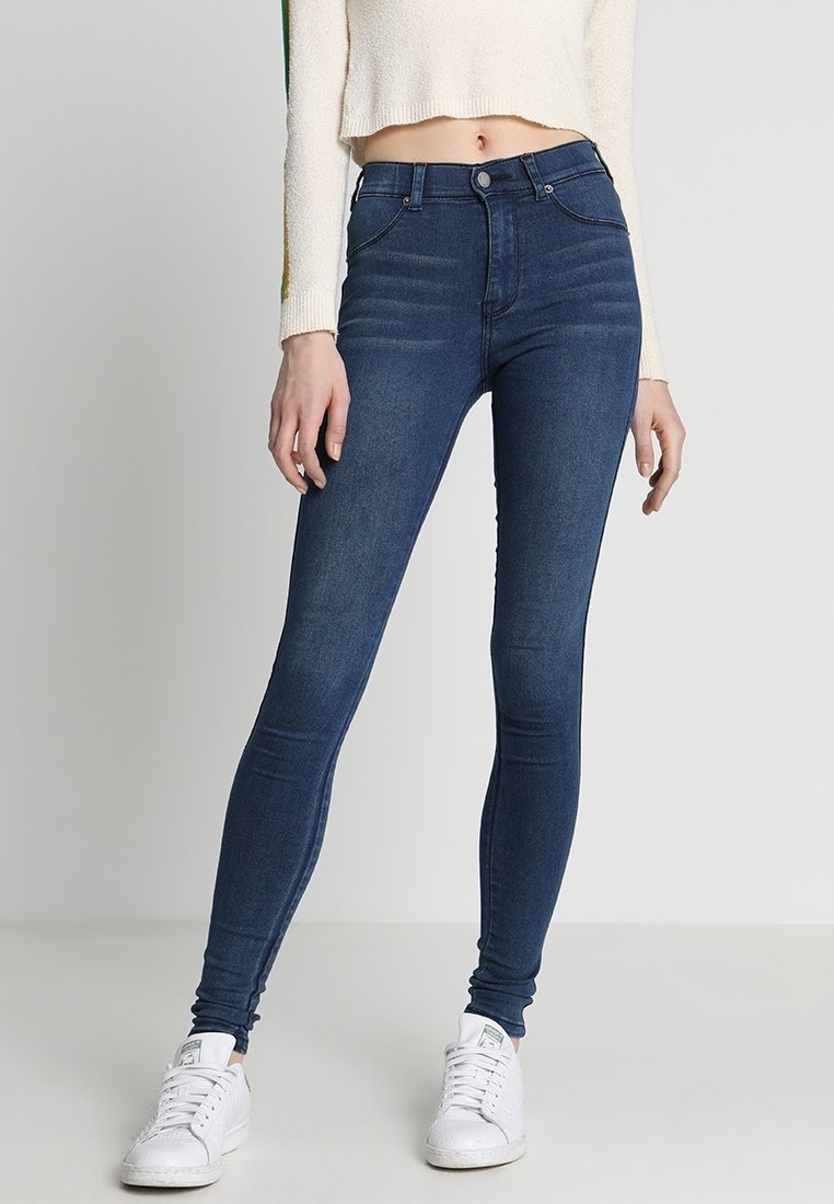 Femme portant un jean skinny bleu foncé, un pull court blanc et des baskets blanches, debout devant un fond clair uni.