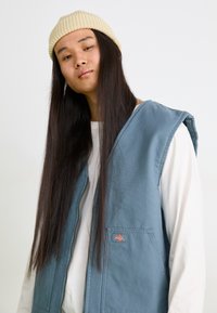 Gilet bleu avec une surface texturée et de larges emmanchures ; porté par-dessus une chemise blanche à manches longues. Comprend un petit patch logo sur la poitrine.