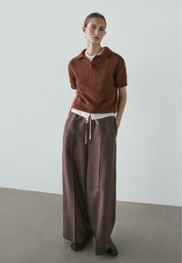 Brązowy sweter dziany w stylu polo z krótkim rękawem, w połączeniu z szerokimi brązowymi spodniami z satyny z wiązanym paskiem w talii i kontrastującym białym akcentem.