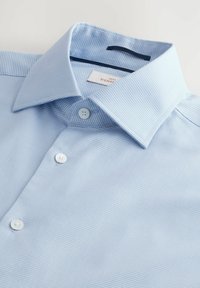Cămașă bleu deschis, cu model texturat, având guler clasic, trei butoni și etichetă în interiorul gulerului.