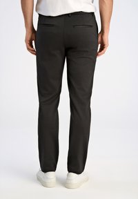 Zwarte broek met een slim fit, voorzien van twee achterzakken met platte sluitingen en een gladde textuur. Gecombineerd met witte sneakers.