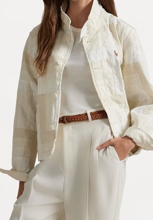 Frau trägt eine cremefarbene Patchwork-Jacke über einem weißen, gerippten Top mit hoch taillierter weißer Hose und braunem geflochtenem Gürtel, Hand in der Tasche.