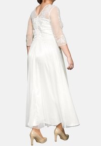 Lång vit klänning med genomskinliga trekvartsärmar, spetsdetaljer på ryggen och en flödande satängskjol. Matchat med nude-hälar.