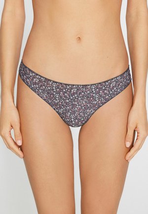 Mujer con braguita de bikini de tiro bajo con un estampado floral oscuro, de pie con las manos relajadas a los lados frente a un fondo sencillo.