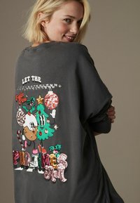 Grauer übergroßer Pullover mit einem bunten, glitzernden Grafik von Cartoonfiguren, Weihnachtsschmuck und verspieltem Text auf der Rückseite.