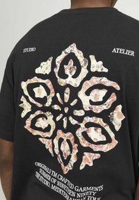 Zwarte t-shirt met een groot abstract bloempatroon in gedempte kleuren op de achterkant. Tekst die verband houdt met het merk en de collectie.