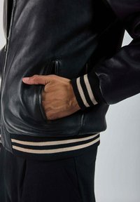 Veste bomber en cuir noir avec fermeture éclair à l'avant. Dotée de poignets et d'une taille en tricot rayé noir et crème. Poche latérale visible.