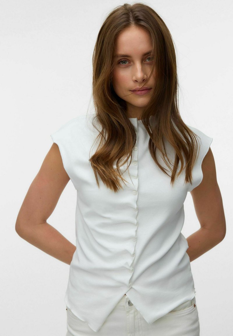 Femme aux longs cheveux bruns portant un haut blanc sans manches boutonné, debout les mains derrière le dos sur un fond clair uni.