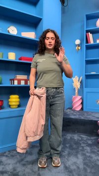 Göndör hajú nő zöld pólóban és farmerben rózsaszín dzsekit tart kék szobában, polcokkal, könyvekkel és dísztárgyakkal.