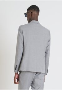 Giacca di suit grigio chiaro su misura con tessuto testurizzato, con spacco singolo e dettaglio a quattro bottoni sui polsini. Indossata con pantaloni abbinati e occhiali da sole.