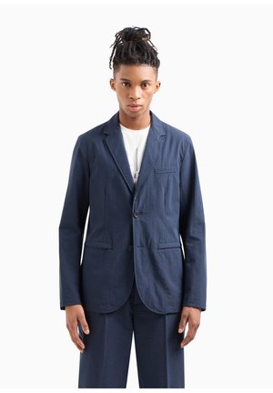 Blazer blu navy con texture, due bottoni, taschino sul petto e tasche a pattina frontali. Il tessuto presenta un motivo sottile ed è di taglio su misura.
