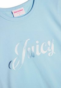 Világoskék, puha anyagból készült póló, fényes ezüst színű "Juicy" logóval, amely dőlt betűkkel van írva, kerek nyakkivágással és rövid ujjakkal.