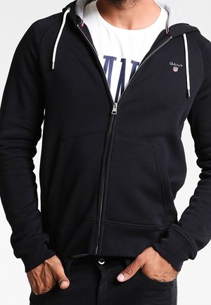 Hoodie noir zippé avec doublure intérieure grise, poches kangourou et poignets côtelés. Présente un petit logo sur la poitrine. Tissu doux et texturé.