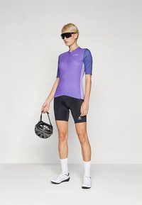 Fialový cyklistický dres s krátkými rukávy, doplněný černými kraťasy. Model drží černou helmu, má na sobě sluneční brýle a bílé cyklistické boty.