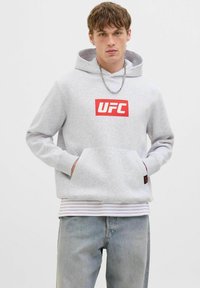 Junger Mann, der einen hellgrauen UFC-Hoodie, eine Kettenhalskette, ein gestreiftes T-Shirt und verwaschene blaue Jeans trägt, steht vor einem schlichten weißen Hintergrund.