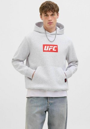 Ung mand iført en lys grå UFC hoodie, kædereg, stribet t-shirt og falmede blå jeans, stående foran en ensfarvet hvid baggrund.