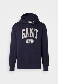 Marineblå hettegenser laget av bomullsblanding. Har en frontlomme i kengurustil, teksturert hvit trykk "GANT 49," og hette med snøring.