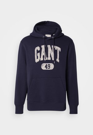 Marineblå hettegenser laget av bomullsblanding. Har en frontlomme i kengurustil, teksturert hvit trykk "GANT 49," og hette med snøring.