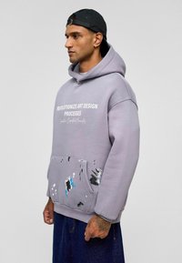 Hoodie van lichtgrijze sweatstof met een voorzak. Bevat artistieke verfspatten in blauw en zwart. Tekst op de borst.