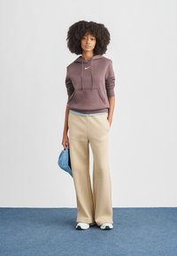 Barna kapucnis pulóver fehér logóval, bézs bő szárú melegítőnadrág, és fehér cipők zöld részletekkel. A modell egy kék sapkát tart. Simább anyagok textúrái.