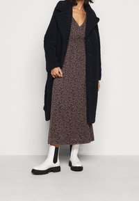 Schwarzer Fleece-Mantel mit großen Revers über einem Midi-Kleid mit Blumenmuster. Das Outfit wird mit weißen Stiefeletten mit schwarzen Sohlen und Akzenten abgerundet.