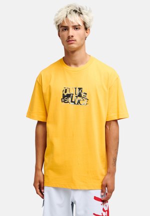 Jeune homme aux cheveux blonds clairs portant un t-shirt graphique jaune et un short blanc avec des marques rouges, debout devant un fond uni.