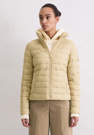 Hellbeige Steppjacke mit vertikalen Nähten, Reißverschluss und Kapuze. Dazu wird ein cremefarbener Strickpullover getragen, kombiniert mit beigen Hosen.