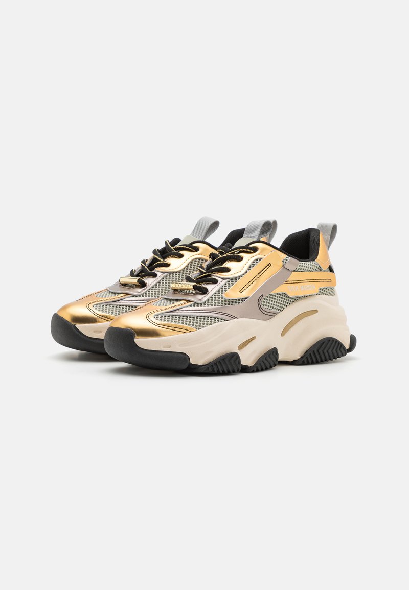Zapatillas con una parte superior de malla gris y dorada metálica, cordones negros y una suela gruesa blanca y negra. Presentan un cuello acolchado y un detalle de logo.