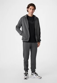 Gråt atletik sæt med lynlåslukket jakke og matchende bukser, med en sort logot-shirt under. Sneakers med sorte og hvide accenter.