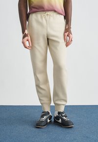 Μπεζ φόρμες με ελαστική μέση και κορδόνι, με στενές μπατζάκια και ριμπ μανσέτες, σε συνδυασμό με μαύρα αθλητικά παπούτσια Nike.