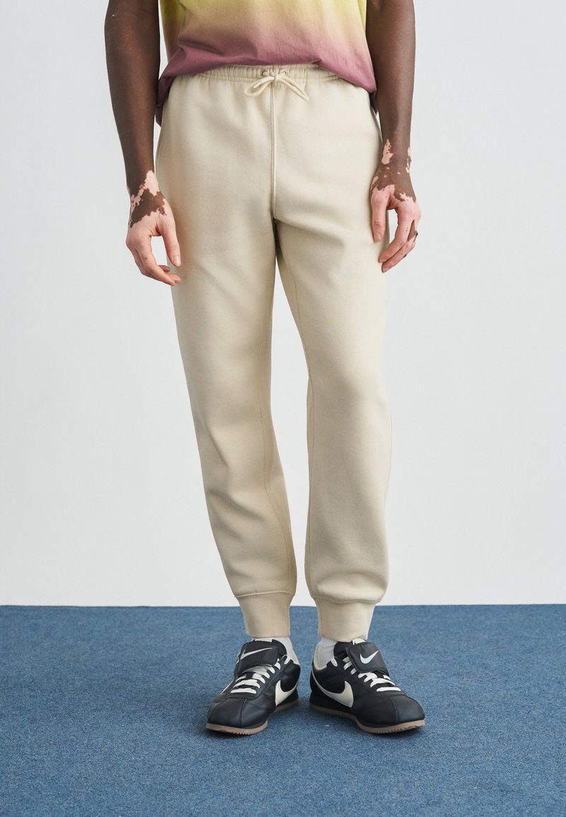 Μπεζ φόρμες με ελαστική μέση και κορδόνι, με στενές μπατζάκια και ριμπ μανσέτες, σε συνδυασμό με μαύρα αθλητικά παπούτσια Nike.