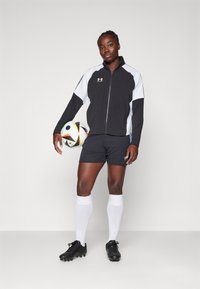 Melna un balta sporta jaka, pelēkas šortus, baltas līdz ceļiem zeķes, melnas futbola kurpes, turot daudzkrāsu futbola bumbu, stāvoša poza.
