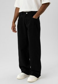 Zwarte wijde broek van zachte stof, voorzien van een hoge taille, steekzakken aan de voorkant en een klassieke knoopsluiting. Gecombineerd met witte sneakers.