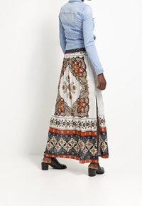 Long, white skirt featuring ornate patterns in orange, blue, and brown, with a side slit. Paired with a light blue denim jacket and black block heels.
