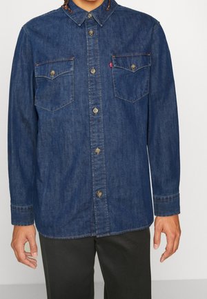 Mies, joka pukeutunut tummansiniseen pitkähihainen farkkupaitaan, jossa on napilliset rintataskut, ja mustiin housuihin, seisoo yksivärisen taustan edessä.